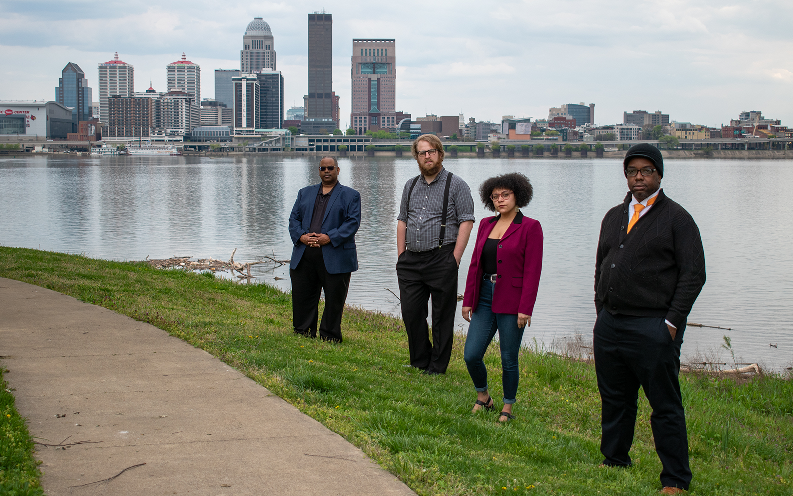 four musicians stand in front of a river and the Louisville Kentucky skyline