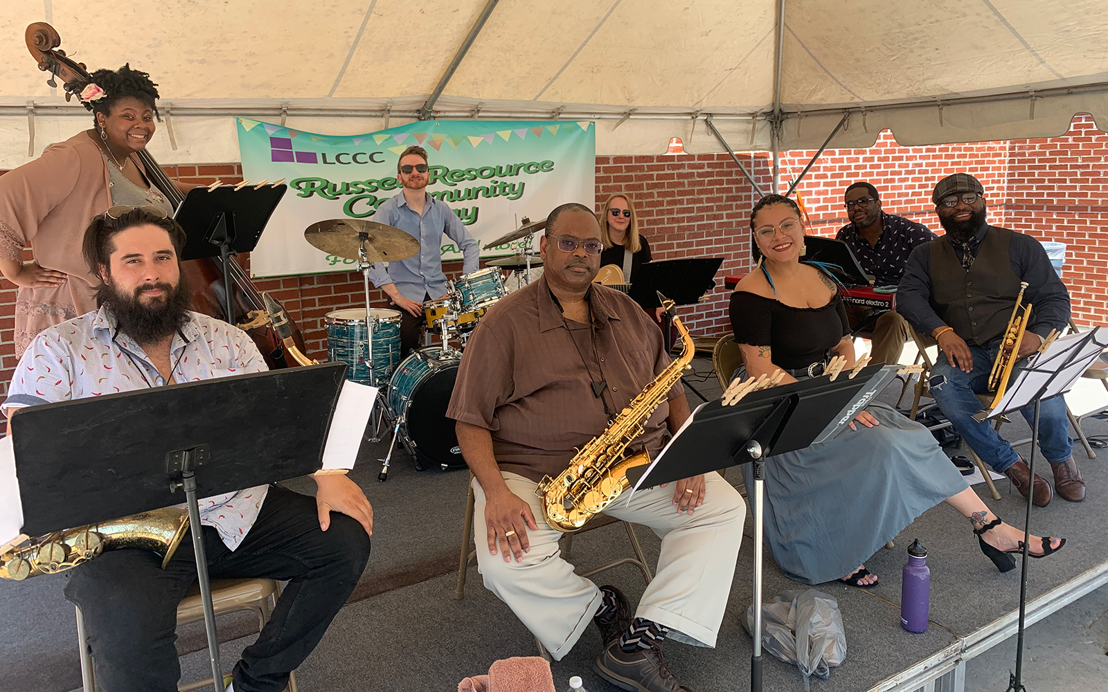 a diverse group of eight musicians with their instruments on a stage; a banner behind them reads "LCCC: Russell Resource Community Day"